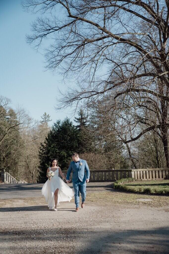 das Brautpaar geht über die Terrasse vom Schloss Wolfsbrunnen und ihr Kleid weht im Wind
