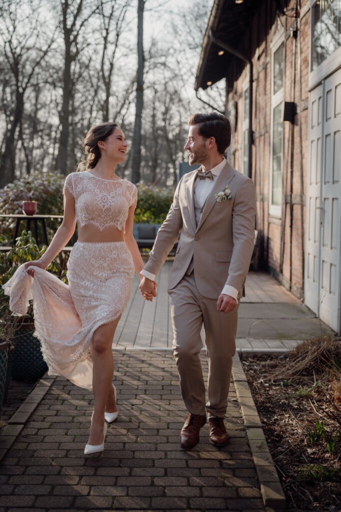 Brautpaar geht gemeinsam an dem Peak Hotel entlang und lächelt sich an, während sie ihr Kleid hoch hält