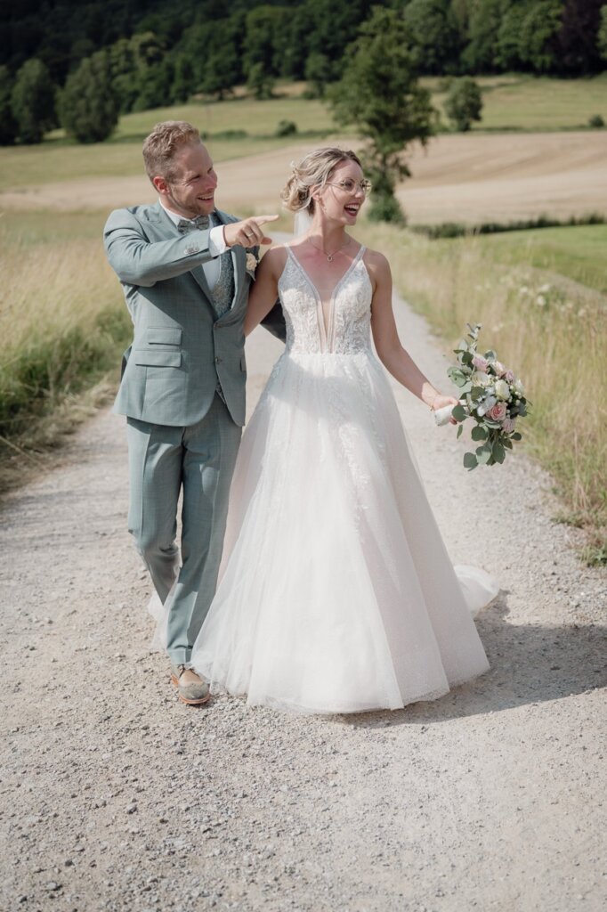 Brautpaar geht durchs Feld und der Brautigam zeigt seiner Braut einen schönen Punkt